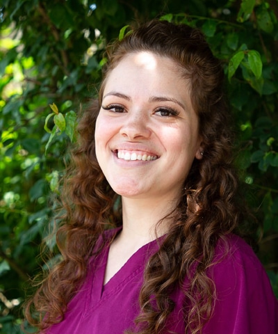 A woman with long curly brown hair wearing a purple top smiles while standing in front of green leafy bushes, representing the Professional Veterinary Expert Team in Rockaway and Denville. Sunlight creates soft highlights on her face and in the background.