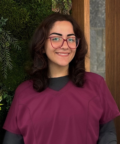 A smiling woman with dark hair and glasses, part of the Professional Veterinary Expert Team in Rockaway and Denville NJ, stands indoors near a wall with green plants and wood paneling, wearing a burgundy scrub top.
