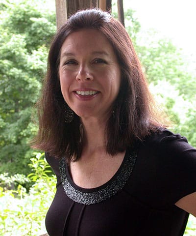 A woman with shoulder-length dark hair, wearing a black top with a sparkly neckline, smiles while standing outdoors in front of green trees—representing the Professional Veterinary Expert Team in Rockaway and Denville.