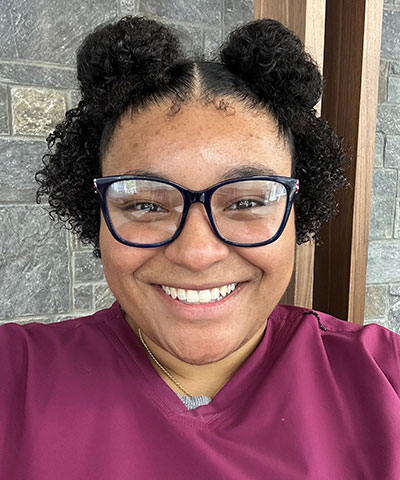 A smiling person with curly hair in two buns, wearing glasses and a maroon shirt, stands in front of a stone and wood wall, representing the Professional Veterinary Expert Team in Denville NJ.