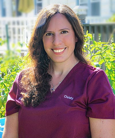 A woman with long brown hair, wearing a maroon scrub top embroidered with "Christy," smiles while standing outdoors in front of green plants and a sunlit fence—part of the Professional Veterinary Expert Team in Rockaway and Denville.