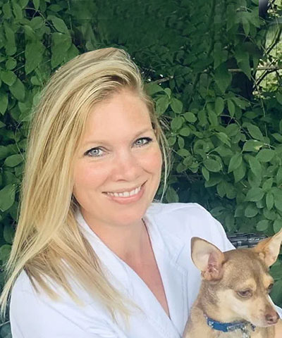 A woman with long blonde hair, wearing a white top, smiles while holding a small brown dog. Green leafy bushes are in the background, reflecting the care and warmth of the Professional Veterinary Expert Team in Denville NJ.