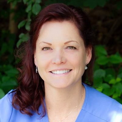 A woman with wavy auburn hair and light skin smiles at the camera. She is wearing a light blue top and small earrings, with green foliage in the blurred background.