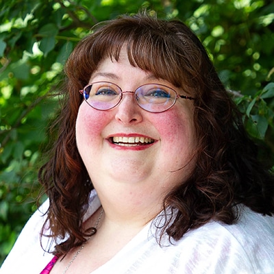 A woman with wavy brown hair and glasses smiles warmly at the camera. She is wearing a white top and is standing outdoors with green foliage in the background.