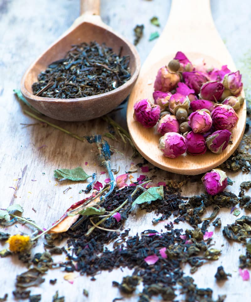 Two wooden spoons on a rustic surface, one filled with loose black tea leaves and the other with small dried pink rosebuds—symbols of Herbal Therapy for Pets in Rockaway and Denville, surrounded by scattered tea leaves and rosebuds.