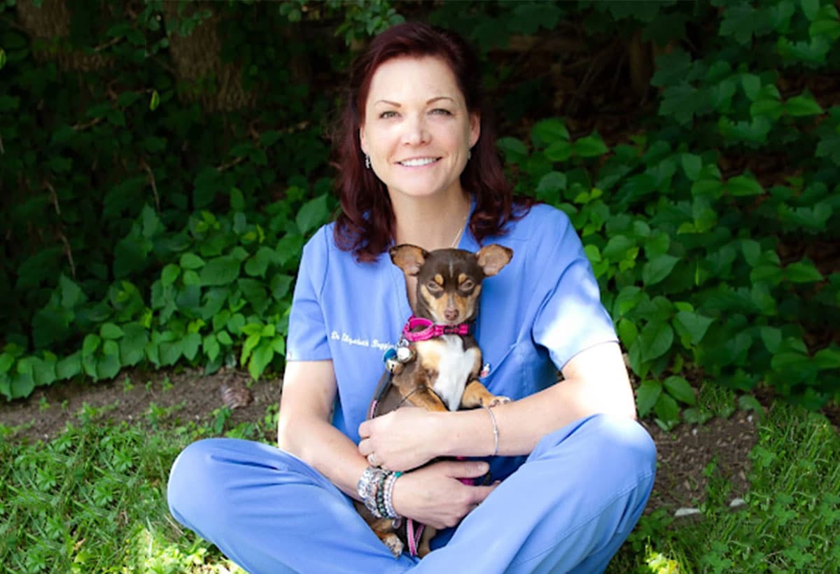 A woman in blue scrubs sits on grass, smiling and holding a small brown and tan dog with a pink collar in her lap. Green foliage and trees are in the background.