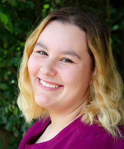 A young person with shoulder-length wavy blonde hair smiles warmly at the camera, wearing a magenta top. Green foliage is blurred in the background—reflecting the friendly atmosphere of our Professional Veterinary Expert Team in Rockaway and Denville.