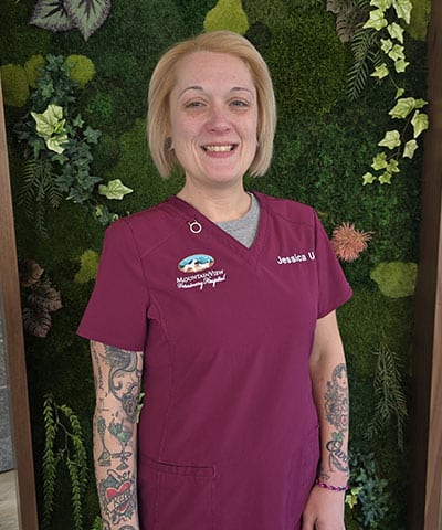 A woman with blonde hair, wearing a maroon scrub top labeled "Jessica U" and a veterinary logo, stands smiling in front of a green plant wall. As part of the Professional Veterinary Expert Team in Denville NJ, she has visible tattoos on both arms.