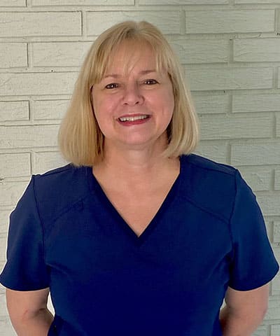 A woman with straight blonde hair wearing a navy blue top stands smiling in front of a white brick wall, representing the Professional Veterinary Expert Team in Rockaway and Denville.