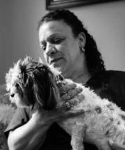 Black and white photo of a woman with curly hair, indoors, gently petting a small dog in her lap. Her caring expression reflects the compassion found in our Professional Veterinary Expert Team in Rockaway and Denville.