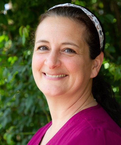 A woman with dark hair pulled back by a patterned headband smiles at the camera in a magenta top. She stands outdoors among green foliage, representing the Professional Veterinary Expert Team in Rockaway and Denville.