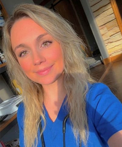 A woman with long blonde hair wearing blue medical scrubs and a stethoscope smiles at the camera in a sunlit room, representing our professional veterinary expert team in Rockaway and Denville NJ.