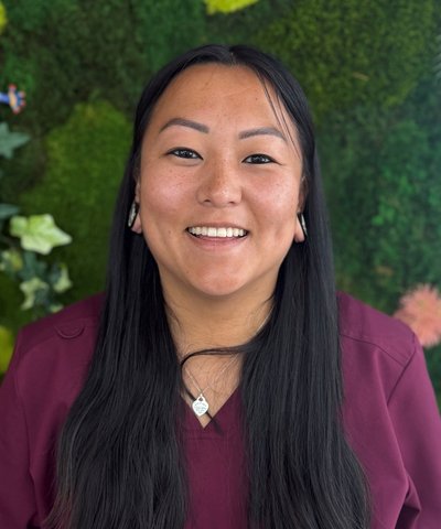 A smiling woman with long dark hair, wearing a maroon scrub top, stands in front of a green plant wall. Liz M is a Customer Service Representative at MountainView Veterinary Hospital in Denville NJ.