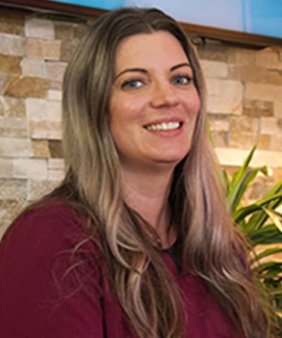 A woman with long, wavy light brown hair smiles at the camera. She is wearing a maroon top and stands in front of a stone wall with green plants, representing the Professional Veterinary Expert Team in Rockaway and Denville.