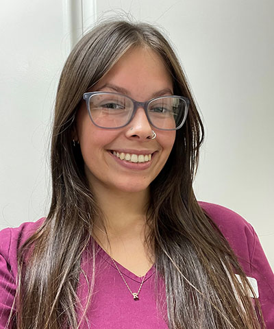 A young woman with long brown hair and glasses smiles at the camera. Wearing a burgundy top and an "R" pendant, she stands against a white background, representing the Professional Veterinary Expert Team in Rockaway and Denville.