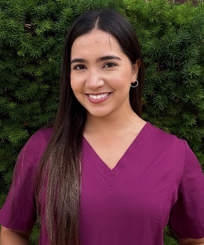 A young woman with long dark hair, wearing a magenta V-neck scrub top, stands smiling in front of green foliage.