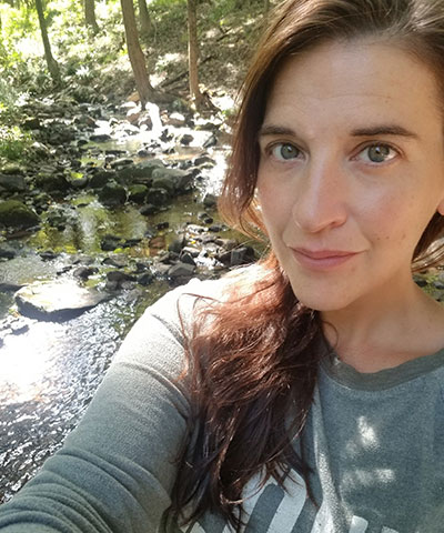 A woman with long brown hair takes a selfie in front of a forest stream, sunlight filtering through the trees. She wears a gray top and slight smile, reminiscent of the friendly professionalism found in our Veterinary Expert Team in Rockaway and Denville.