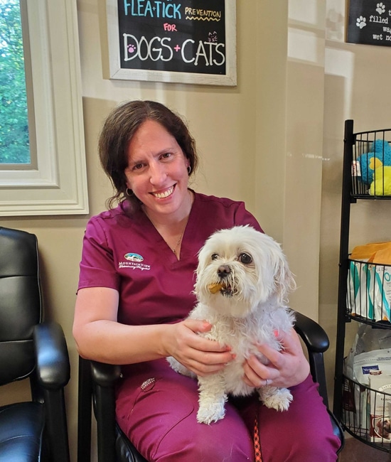 A woman in maroon scrubs sits in a chair, smiling while holding a small white dog. Behind her, a sign about flea and tick prevention for dogs and cats is on the wall.