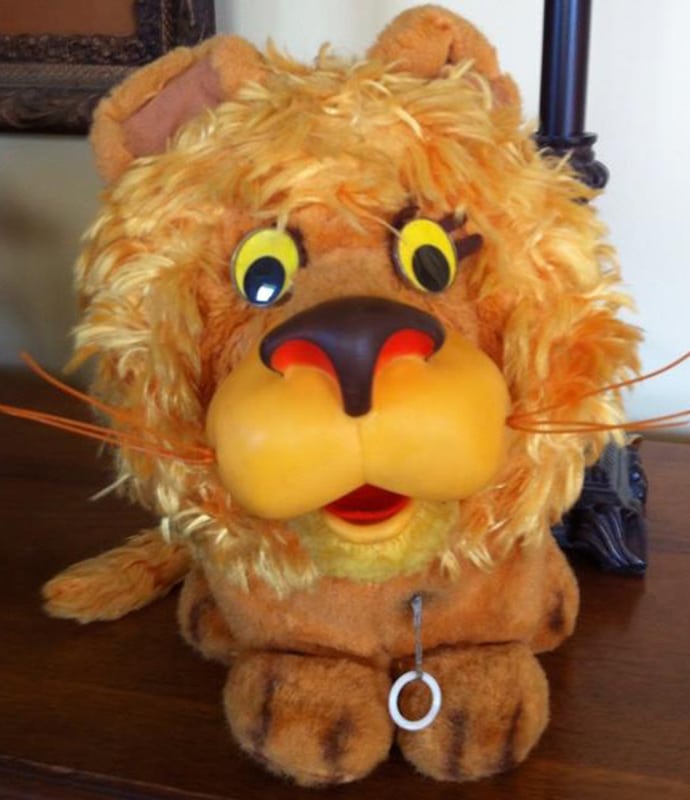 A plush toy lion with a fluffy mane, big yellow eyes, a large nose, and a pull string on its chest sits on a wooden surface. The lion has a cartoonish, friendly expression.