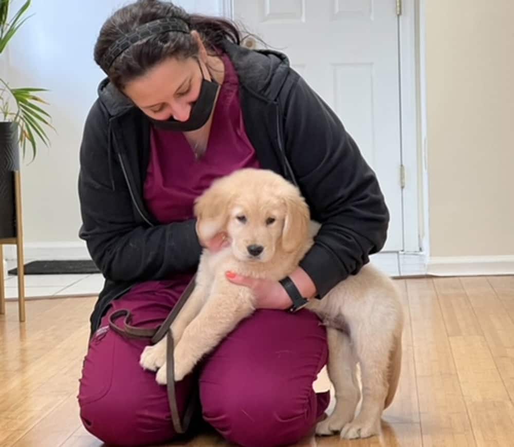 A woman in burgundy scrubs and a black mask kneels on the floor, gently hugging a fluffy golden retriever puppy indoors near a white door.