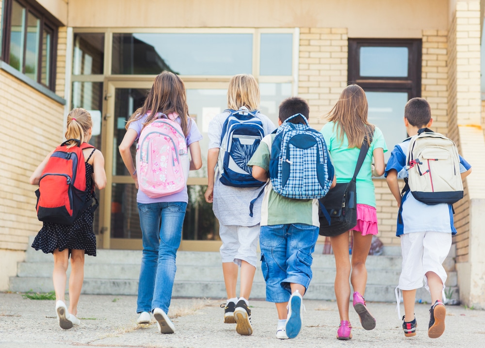 Six children with backpacks walk together toward the entrance of a school building, seen from behind. The brick building has large windows and two doors. The kids appear to be heading to class.